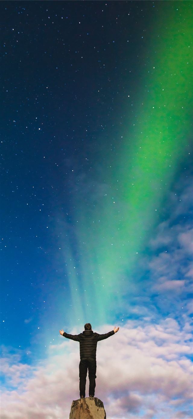 man standing on rock under green aurora borealis a... iPhone 12 wallpaper 