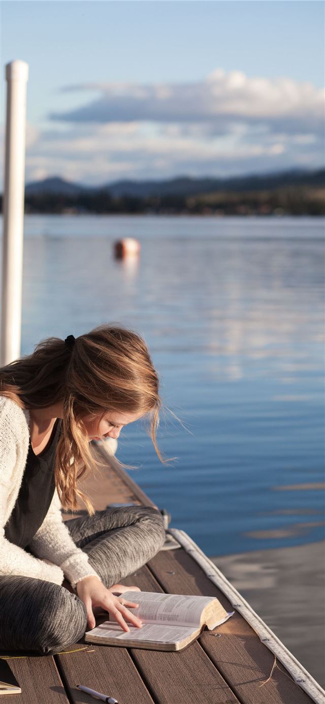 photo of woman reading book near body of water iPhone 12 wallpaper 
