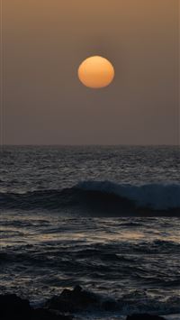 A large orange sun setting over a dark choppy ocea... wallpaper