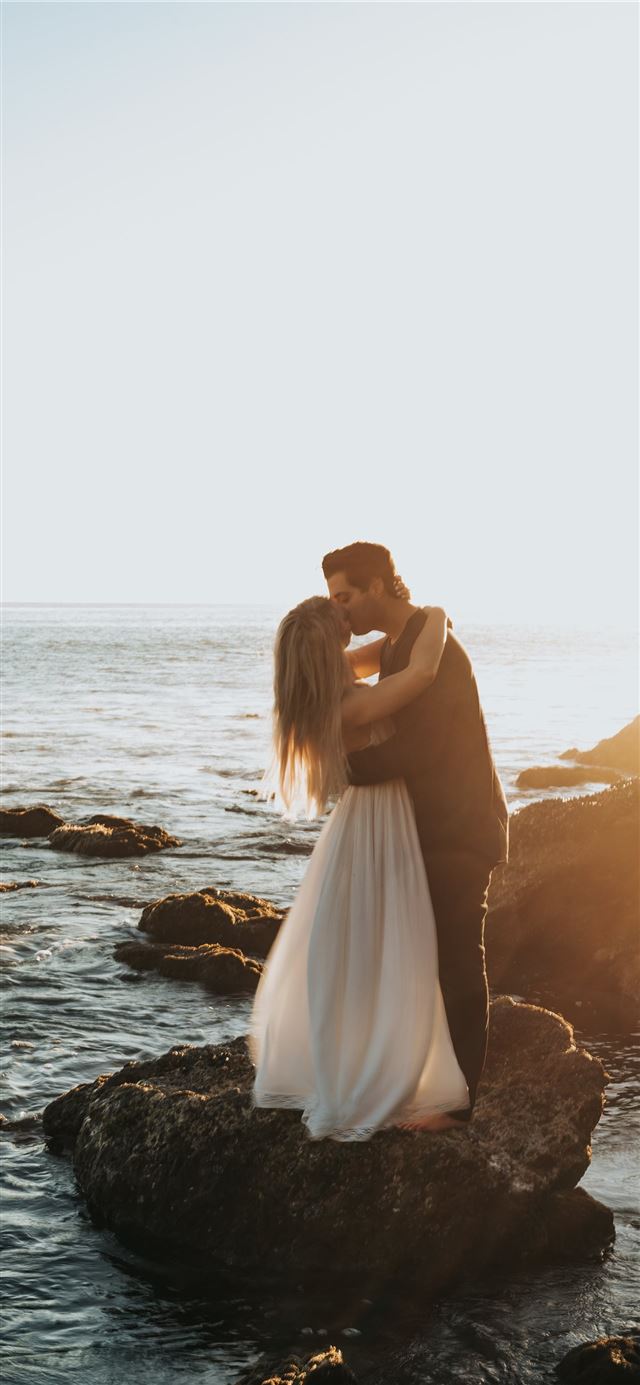 man and woman kissing on top of gray rock at beach iPhone 11 wallpaper 